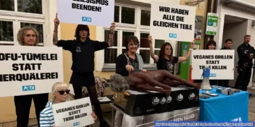PETA Schweinefest wird zum Desaster – Wenn Aktivismus nach Räucherspeck riecht 46 Sechs PETA-Aktivisten stehen mit traurigen Gesichtern an einem trostlosen Infostand beim PETA Schweinefest in Viechtach, während im Hintergrund fröhlich gegrillt wird.