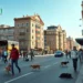 Das Bild zeigt eine lebhafte Straßenszene in Istanbul an einem sonnigen Tag. Menschen eilen geschäftig durch die Straßen, umgeben von einer Mischung aus modernen und traditionellen Gebäuden. Inmitten der Stadtaktivität sind mehrere streunende Hunde und Katzen zu sehen, die entweder umherstreifen oder sich ausruhen. Plakate und Schilder im Hintergrund thematisieren subtil den Tierschutz und appellieren an das Bewusstsein für das Wohlergehen der Tiere. Die Komposition des Bildes fängt die Dynamik des Stadtlebens ein, während die Präsenz der Tiere die Dringlichkeit der Tierschutzdebatte unterstreicht.