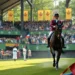 Das Titelbild zeigt eine realistische Darstellung des Concours Hippique International Officiel (CHIO) in Aachen, einem renommierten Reitsport-Event. Im Vordergrund reitet ein eleganter Reiter sein Pferd geschickt durch einen anspruchsvollen Parcours, der die Tradition und Anmut des Reitsports verkörpert. Im Hintergrund ist eine lebhafte Zuschauermenge zu sehen, einige halten Flugblätter in der Hand, was auf die aktuelle Debatte zwischen Tradition und Tierschutzaktivismus hinweist. Die üppige grüne Kulisse der Arena ist mit Bannern und Dekorationen geschmückt, die das kulturelle Erbe des Events betonen. Die Atmosphäre vermittelt sowohl die Leidenschaft für den Reitsport als auch die unterschwellige Spannung der Kontroverse. Kleine Gruppen von Menschen, die in Diskussionen vertieft sind, symbolisieren den Konflikt zwischen traditionellem Reitsport und modernen Tierrechtsbewegungen.