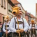 Ein fotorealistisches Bild zeigt eine traditionelle deutsche Schützenfest-Parade ohne Pferde. Menschen in Trachten ziehen stolz durch das historische Stadtzentrum, das von charmanten alten Gebäuden gesäumt ist. Die festliche Atmosphäre wird durch bunte Fahnen und traditionelle Musikkapellen untermalt, während fröhliche Menschenmengen die gepflasterten Straßen säumen. Im Hintergrund sind Anzeichen von üppigem Grün und entfernten, sanften Hügeln zu sehen, die die Schönheit der deutschen Landschaft andeuten. Das Bild vermittelt ein Gefühl von kulturellem Erbe und Gemeinschaftsgeist und zeigt, wie sich Traditionen in einem modernen Kontext weiterentwickeln, auch ohne die früher typischen Pferde bei Schützenfesten.
