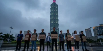 PETA - Alex Honnold und die Inszenierung veganer Leistungsfähigkeit 46 Aktivisten stehen mit leeren Schildern vor dem beleuchteten Taipei 101 bei Regen, während hoch oben ein Kletterer zu sehen ist und die Szene die umstrittene Aktion PETA Alex Honnold symbolisiert